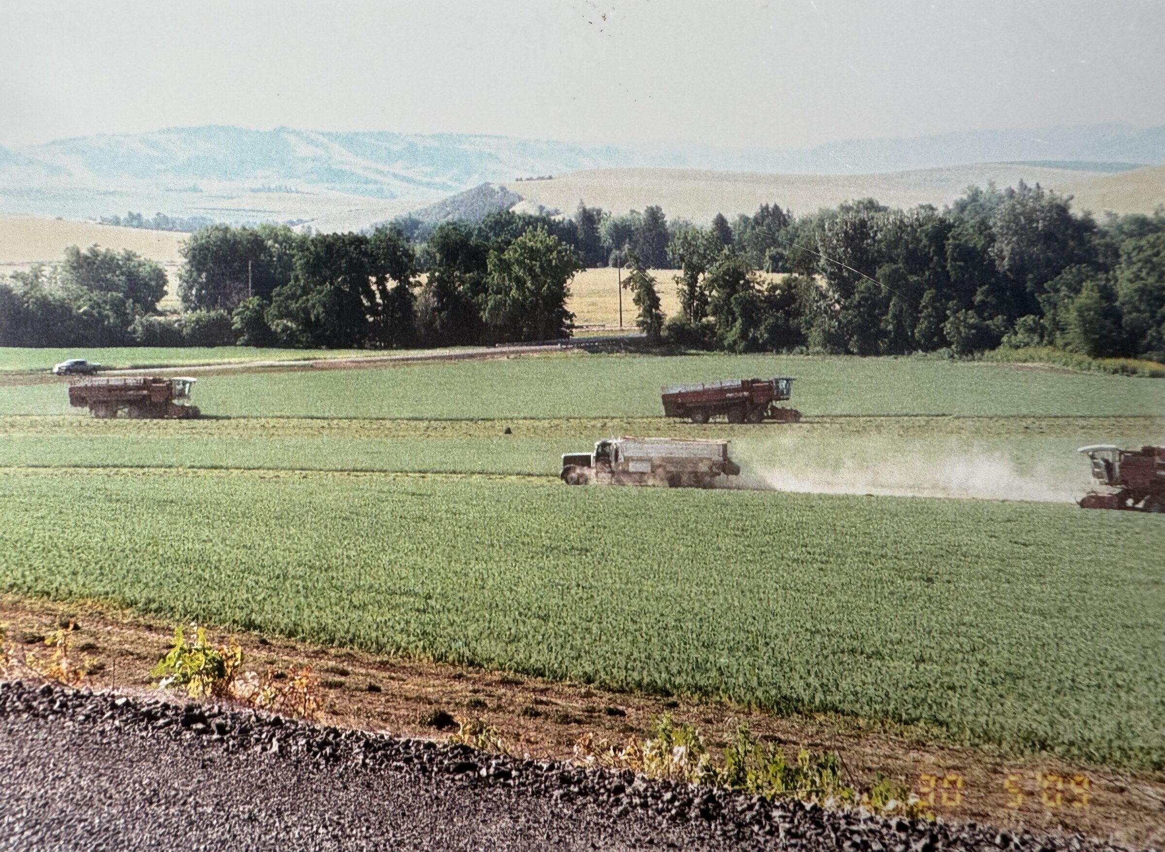 Trucks and combines working a green field below rolling hills